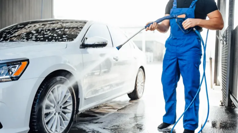 Muž v modrých kalhotách umývá bílé auto vysokotlakou myčkou.