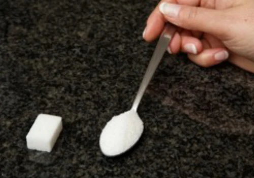 Jedna kostka cukru vedle lžičky granulovaného cukru na tmavém povrchu.