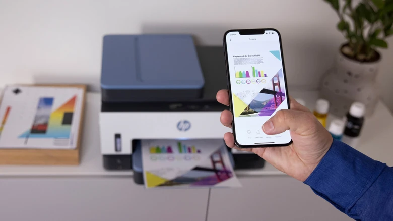 Ruka drží chytrý telefon s aplikací pro tiskárnu HP vedle funkční bezdrátové tiskárny.