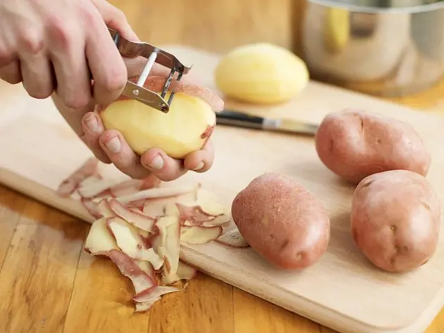 Člověk škrábe kůru z brambory pomocí ošklivky na dřevěném prknu.