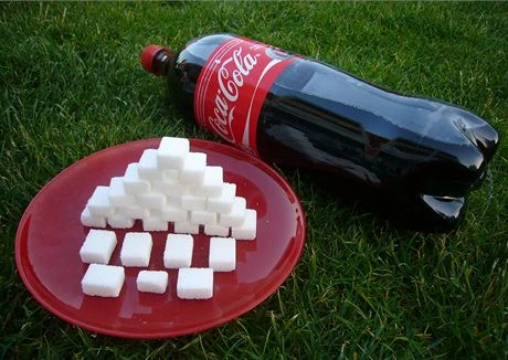 Sklenice Coca-Coly vedle talíře s kostkami cukru na trávě.