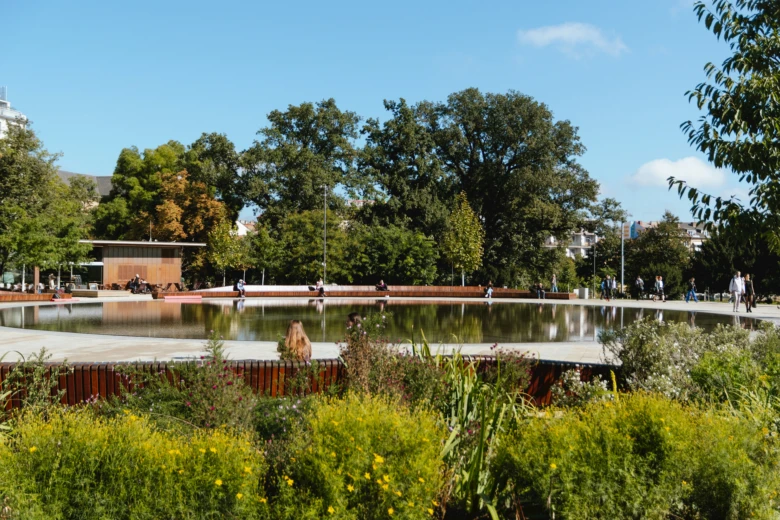 Sluneční městský park s vodní plochou, stromy a lidmi.