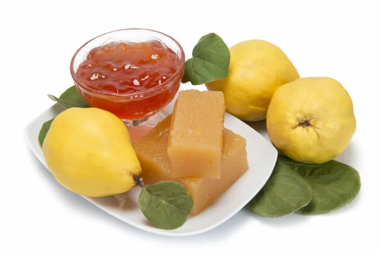 Žluté kdoule s lístky na bílém talíři vedle marmelády.