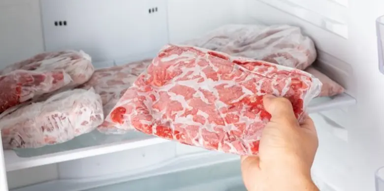 Ruka drží balení surového masa v mrazáku s dalšími balíčky.
