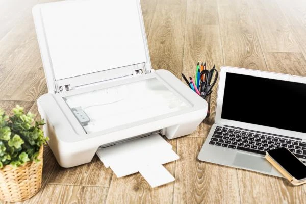 Bílá tiskárna na dřevěné podlaze vedle notebooku s tužkami.