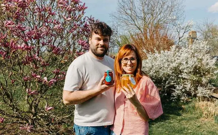Muž a žena drží sklenice medu v zahradě s kvetoucími stromy.