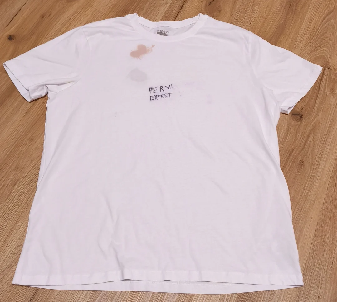 Bílá trička s nálepkou Persil Expert a skvrnami na dřevěném podlaze.