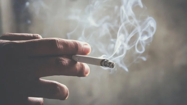 Ruka drží zapálenou cigaretu s kouřem v bytě.