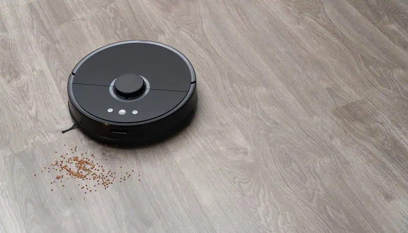 Černý robota vysavač čistí rozsypané krupičky na podlaze.