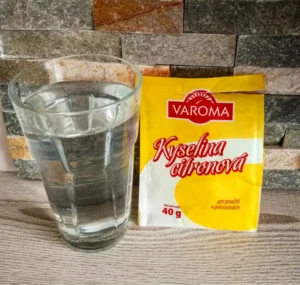 Žlutý balíček kyseliny citronové vedle sklenice vody na dřevě.