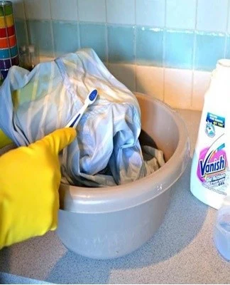 Ruce v gumě čistí skvrnu na oblečení prostředkem Vanish.