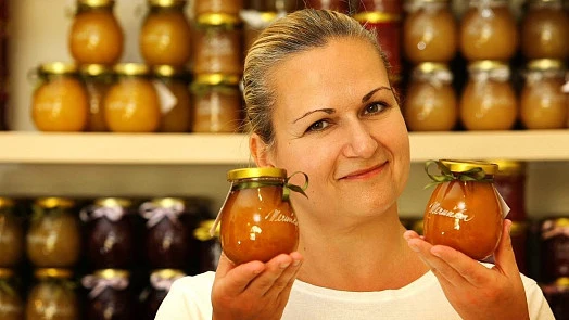 Žena drží dva sklenice s ovocným džemem před regálem plným podobných produktů.
